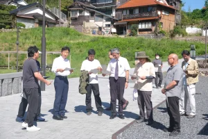 260924nenjitenken1 集合場所の恩湯広場で、資料を手に説明を聞く行政と地域団体の参加者たち