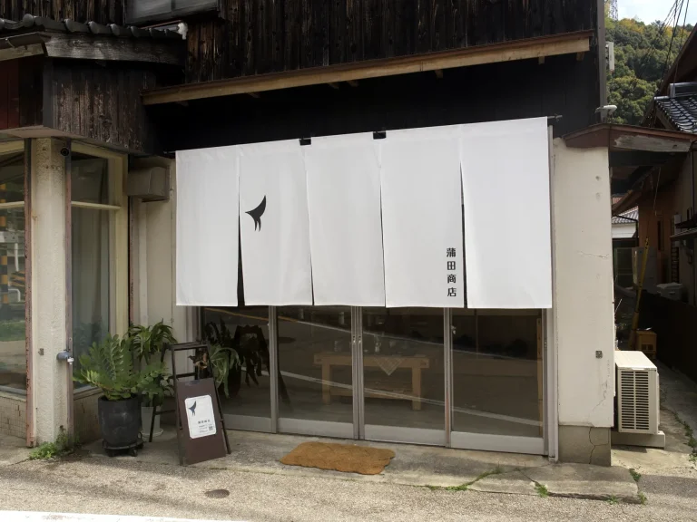 白いのれんに「蒲田商店」の文字、ガラス戸の脇に植木と看板を置いた蒲田商店の店構え