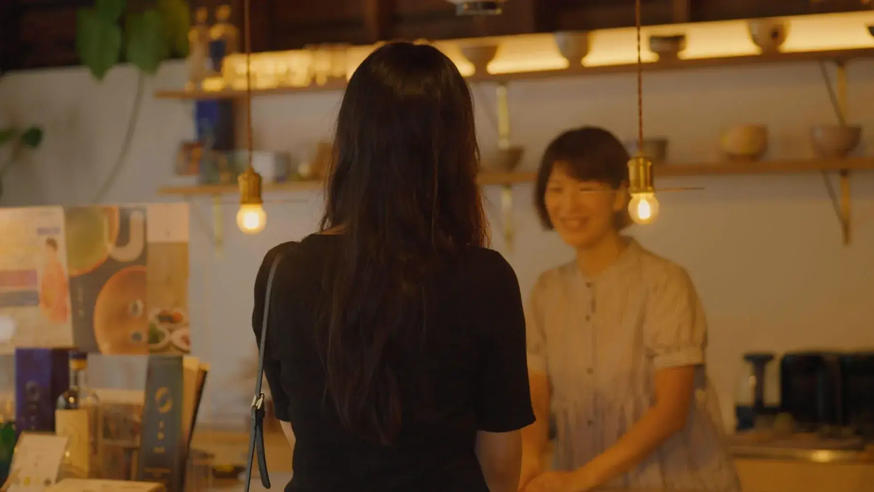 器の並ぶ店で、店の人と向き合って話す女性の後ろ姿
