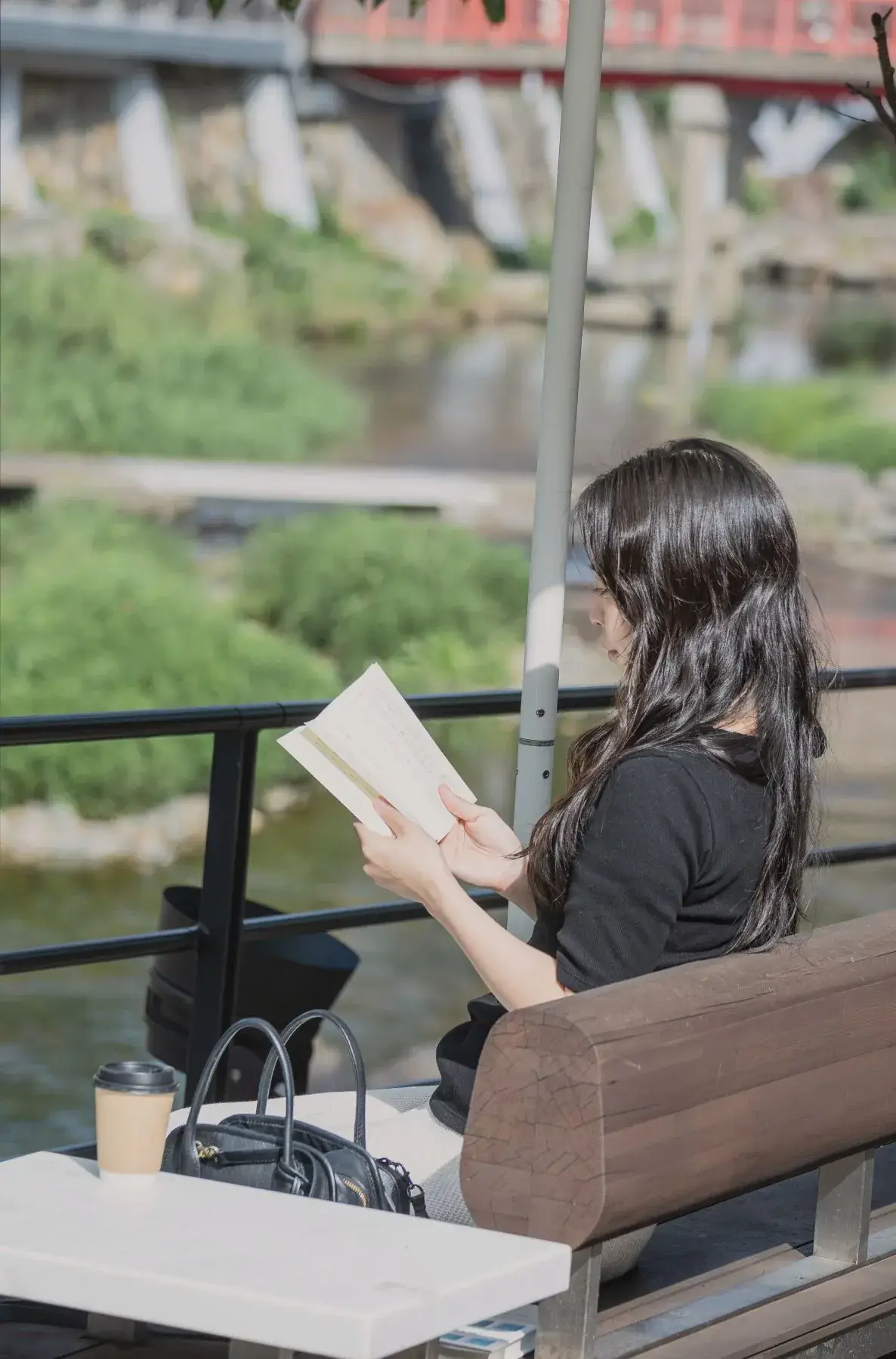 川沿いのベンチでコーヒーを置いて本を読む女性